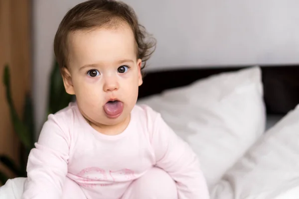 Autentico ritratto carino caucasico bambino paffuto bambina o ragazzo in rosa assonnato al risveglio guardando la fotocamera mostrando la lingua nel letto bianco. infanzia, infanzia, paternità, concetto di stile di vita — Foto Stock