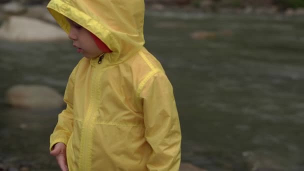 Kleine Vorschulkinder in der Nähe des Bergflusses. Kind in gelbem Regenmantel wirft Stein auf Fluss. Junge warf Pflastersteine vom Ufer ins Wasser. Urlaub, Zeltlager, Frühling, wilde Natur, Familienkonzept