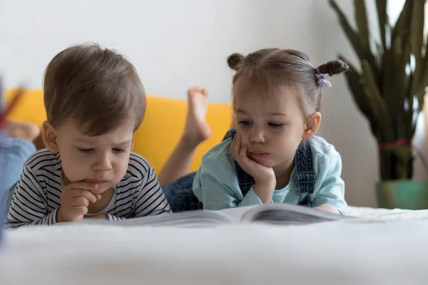 Duas crianças pré-escolares lêem um grande livro interessante de contos de fadas na cama amarela. Os irmãos pequenos leitores de gêmeos se divertem, garoto feliz em quarentena em casa. Amizade, família, conceito de educação — Fotografia de Stock