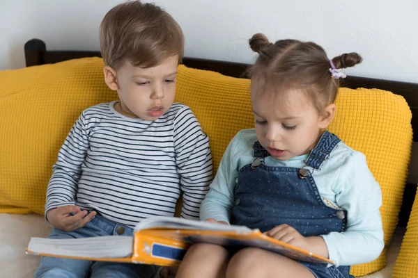 Duas crianças pré-escolares lêem um grande livro interessante de contos de fadas na cama amarela. Os irmãos pequenos leitores de gêmeos se divertem, garoto feliz em quarentena em casa. Amizade, família, conceito de educação — Fotografia de Stock