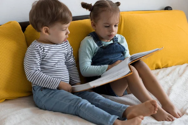 Dos niños preshool, niños pequeños leen gran libro interesante de cuentos de hadas en la cama amarilla. Hermanos pequeños gemelos lector divertirse, niño feliz en cuarentena en casa. Amistad, familia, concepto educativo — Foto de Stock