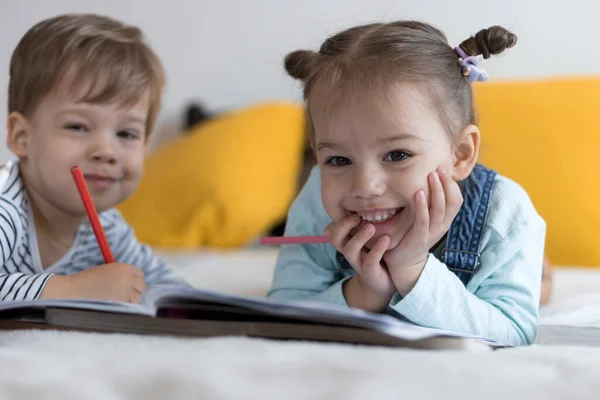 Zeki, gülümseyen, küçük bir çocuk çocuk ve kız sarı yatakta göbeklerinde kalemlerle resim yapıyorlar. Küçük sevimli okuyucu evde karantinada eğleniyor, mutlu çocuk. Arkadaşlık, aile, eğitim. — Stok fotoğraf