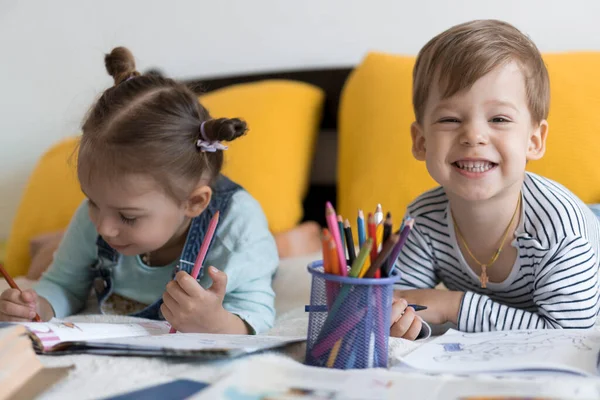 Inteligente sorrindo criança pré-escolar crianças menino e menina desenhar com lápis deitado em seu estômago na cama amarela. leitor bonito pouco se divertir, garoto feliz em quarentena em casa. Amizade, família, educação — Fotografia de Stock