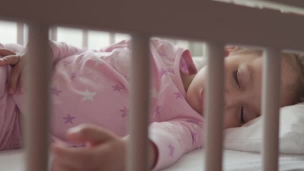 Nahaufnahme authentische niedliche kaukasische kleine Vorschulkind Mädchen schlafen süß in weißen Krippe auf Pyjama im Schlafzimmer während der Mittagszeit. Kind liegt im Bett neben Fenster. Kindheit, Medizin, Gesundheitskonzept — Stockvideo