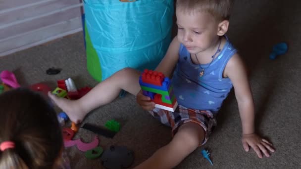 Kleine kleuters en broertjes en zusjes zitten op de vloer met speelgoed in de kinderkamer. De jongens spelen met de bouwvakker in de speelkamer. Kinderen hebben veel plezier thuis gehad. Jeugd, vriendschap, ontwikkelingsconcept — Stockvideo