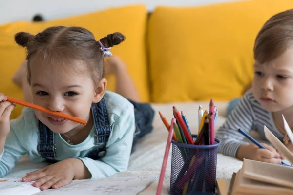 Inteligente sorrindo criança pré-escolar crianças menino e menina desenhar com lápis deitado em seu estômago na cama amarela. leitor bonito pouco se divertir, garoto feliz em quarentena em casa. Amizade, família, educação — Fotografia de Stock