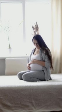 Mutlu hamile kadının evde karnına dokunduğu, şefkatli annenin karnını güneşli bir yaz gününde elleriyle okşadığı dikey format videosu. hamilelik, annelik, insanlar, beklenti kavramı