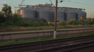 Yüksek hızlı trenin penceresinden eski paslı yük vagonlarının manzarası ve yaz arkaplanındaki güzel bulutlu mavi gökyüzünde çelik tahıl siloları. Ulaşım, seyahat, demiryolu, yol, zina kavramı