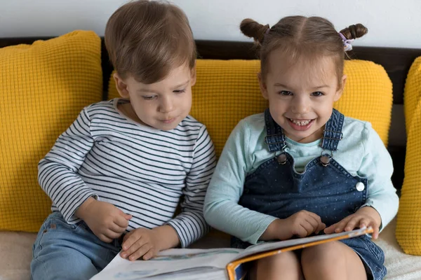 Dvě děti z předškolního batolete čtou na žluté posteli velkou zajímavou knížku pohádek. Sourozenci, dvojčata, čtečka se baví, šťastné dítě v karanténě doma. Přátelství, rodina, koncepce vzdělávání — Stock fotografie