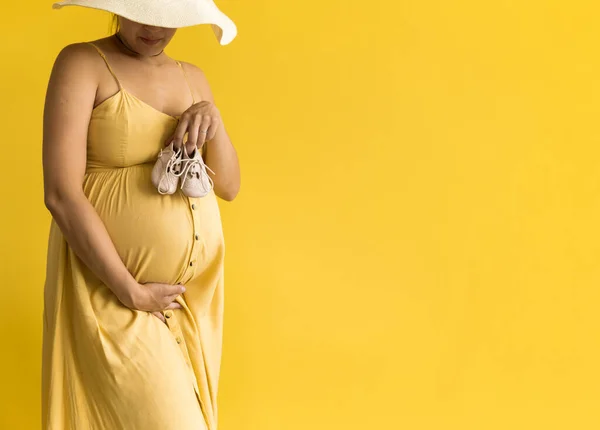 Motherhood, femininity, fashion, hot summer - banner Close-up unrecognizable pregnant woman lady in beautiful vintage dress Provence straw hat with small baby shoes on yellow background, copy space