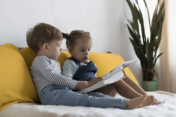 Δύο παιδιά preshool, μικρό παιδί διαβάσει μεγάλο ενδιαφέρον βιβλίο των παραμυθιών στο κίτρινο κρεβάτι. Αδελφές δίδυμες αναγνώστριες διασκεδάζουν, χαρούμενο παιδί σε καραντίνα στο σπίτι. Φιλία, οικογένεια, εκπαίδευση — Φωτογραφία Αρχείου