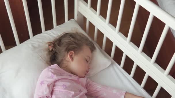 Primer plano auténtico lindo caucásico pequeña niña preescolar dormir dulcemente en cuna blanca en pijama en el dormitorio durante la hora del almuerzo. el chiquitín es el descanso se acuesta cerca de la ventana. Infancia, medicina, concepto de salud — Vídeo de stock