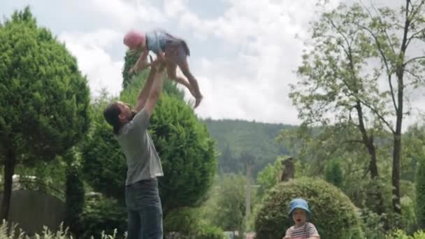 Happy Family Funny Young Dad with Beard Throws the Kids Up High by Arms. Father, Preschool Daughter and Son Have Fun Playing Games in the Backyard Lawn of Idyllic Suburban House on Sunny Summer Day. — Video Stock