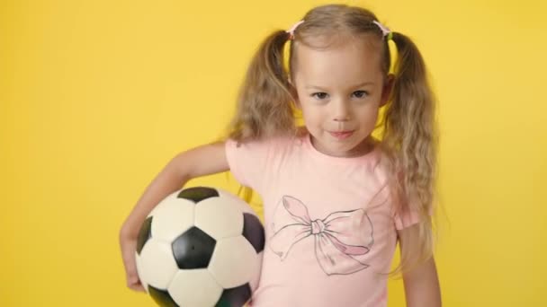 Autentico carino sorridente bambina prescolare con classico pallone da calcio in bianco e nero guardare la fotocamera su sfondo giallo. bambino giocare a calcio in t-shirt e pantaloncini. Sport, campionato, concetto di squadra — Video Stock