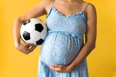 Annelik, kadınlık, futbol, spor, mandıra, sıcak yaz. Kırpılmış, tanımlanamayan, çiçek mavisi elbiseli genç ve güzel bir kadın futbol beyaz ve siyah top sarı arka planda göbeğini okşuyor.