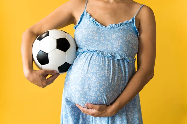 Motherhood, femininity, football, sport, dairy, hot summer. croped unrecognizable pregnant young pretty woman in floral blue dress holds soccer white and black ball rubs tummy on yellow background Stock Image