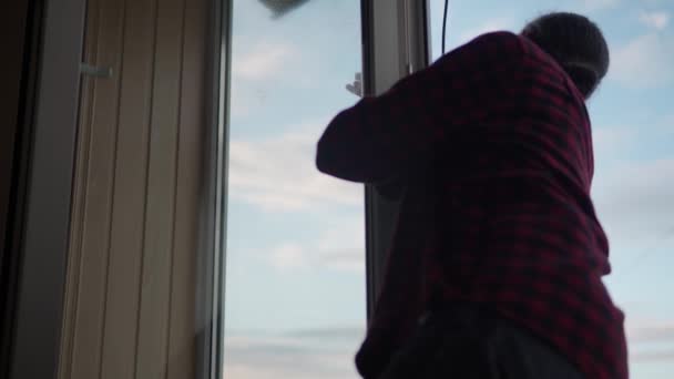 Lavando cristales de ventana en casa. Un joven de pelo largo con barba está limpiando el apartamento. El trabajador sanitario está desinfectando ventanas panorámicas en una habitación vacía. pureza, limpieza, cambio de concepto — Vídeos de Stock