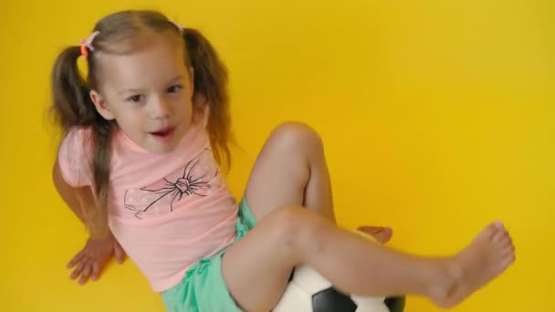 Auténtica linda y sonriente niña preescolar con la clásica bola de fútbol blanco y negro mirar a la cámara en el fondo amarillo. niño jugar al fútbol en camiseta y pantalones cortos. Deporte, campeonato, concepto de equipo — Vídeos de Stock