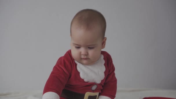 Joyeux noël, bonne année, nourrissons, enfance, concept de vacances - gros plan souriant 7 mois nouveau-né drôle dans santa claus chapeau, costume rouge jouer assis sur lit doux chaud lécher bonbons de Noël doux — Video