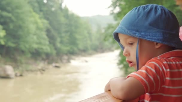 Authentic Little Preschool Cute Caucasian Boy Child in Blue Panama and Red T-shirt Stands on Dais and Looks Into Distance at the Mountain River Дитинство, природа, відпустка, гаряче літо, подорож концепт — стокове відео