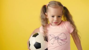 Klasik siyah-beyaz futbol topuyla gülümseyen sevimli anaokulu kızı sarı arka plandaki kameraya bakıyor. Çocuk tişört ve şortla futbol oynuyor. Spor, şampiyonluk, takım konsepti.