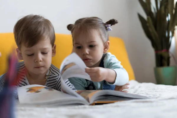Deux enfants d'âge préscolaire, tout-petits lire grand livre intéressant de contes de fées sur lit jaune. Frères et soeurs les petits jumeaux lecteur ont plaisir, enfant heureux en quarantaine à la maison. Amitié, famille, concept d'éducation — Photo