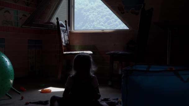 Silhouette kis óvodás lány gyermek ül a földön a játékokat a gyerekek szobában szemben ablak kilátással a hegyekre. A gyerekek sok időt töltöttek otthon. Gyermekkor, barátság, fejlesztési koncepció