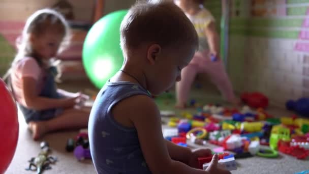 Små förskolebarn som sitter på golvet med leksaker i barnrummet. Killar leker med konstruktörer i lekrummet. Ungarna har spenderat kul hemma. Barndom, vänskap, utvecklingskoncept — Stockvideo