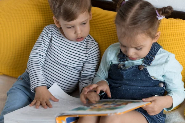 Duas crianças pré-escolares lêem um grande livro interessante de contos de fadas na cama amarela. Os irmãos pequenos leitores de gêmeos se divertem, garoto feliz em quarentena em casa. Amizade, família, conceito de educação — Fotografia de Stock