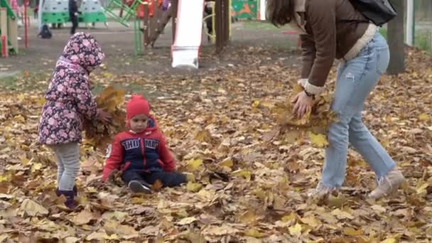 Infanzia, famiglia, neonato, maternità, concetto di autunno - rallentatore 2 allegri bambini piccoli in età prescolare con la mamma raccolgono e vomitano un sacco di foglie gialle cadute vicino al parco giochi nella stagione fredda — Video Stock