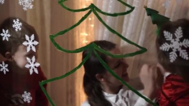 Otantik sevimli bir anne ve iki küçük anaokulu öğrencisi kız ve erkek kardeş 2-4 yaşlarında pencereye Noel ağacı çiziyor. Çocuklu genç bir kadın kar taneleri yapıştırıyor. Xmas, yeni yıl, kış konsepti.