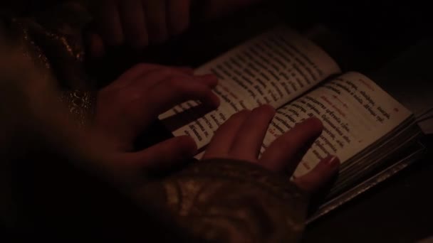 Religia, wakacje, koncepcja - zbliżenie dziecięcych rąk trzymających starożytną książkę i czytających następujący tekst palcem w ciemnym pokoju przy świetle świecy. Słudzy kościelni odprawiają nabożeństwa w kościele — Wideo stockowe