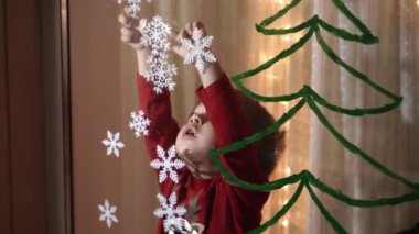 Otantik sevimli bir anne ve iki küçük anaokulu öğrencisi kız ve erkek kardeş 2-4 yaşlarında pencereye Noel ağacı çiziyor. Çocuklu genç bir kadın kar taneleri yapıştırıyor. Xmas, yeni yıl, kış konsepti.