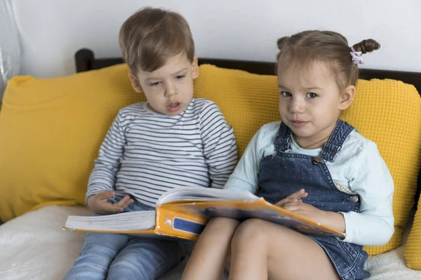 两个早熟、蹒跚学步的孩子在黄色的床上读着一本有趣的童话故事大书。兄弟姐妹的双胞胎读者在家里享受着快乐快乐的孩子被隔离的生活。友谊、家庭、教育概念 — 图库照片