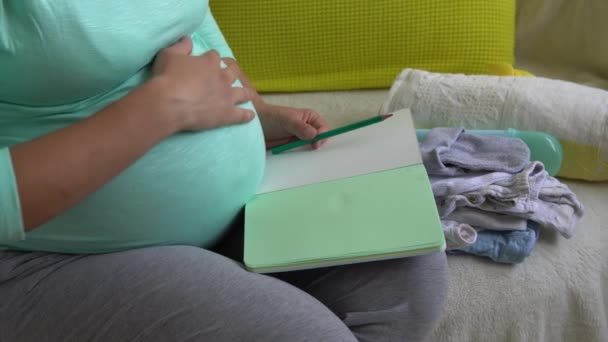 かわいい妊娠中の女性は、ノートブックで妊婦病院のための梱包リストを書く準備袋。妊娠中の若い女性は自宅でソファに楽しい時間を過ごす持っています.母親の健康とケアの概念 — ストック動画