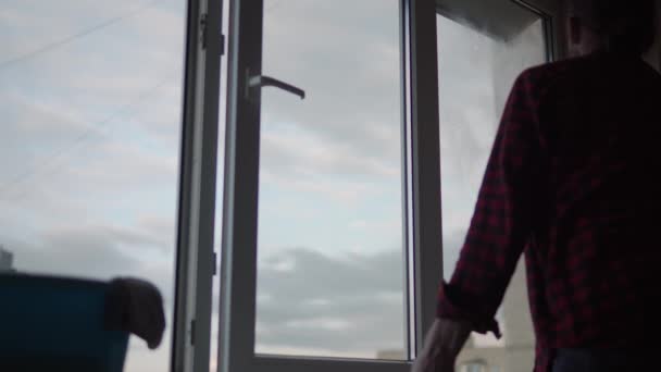 Миття віконного скла вдома. Молодий довгошерстий чоловік з бородою прибирає квартиру. Санітарний працівник дезінфікує панорамні вікна у порожній кімнаті. чистота, чистота, зміна концепції — стокове відео