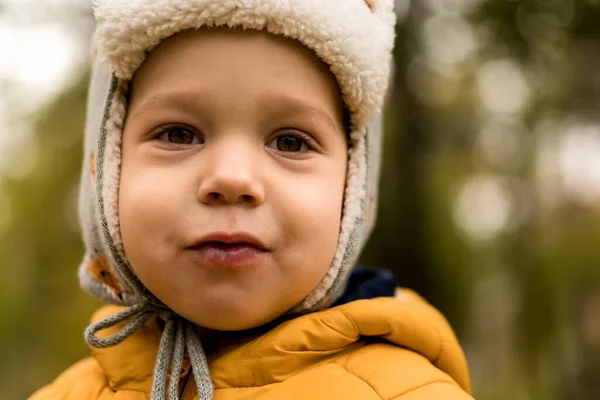 Primer plano Retrato de niño pequeño preescolar lindo en sombrero con solapas de oído y chaqueta naranja Niza sonriente mirando a la cámara en clima frío en el parque de otoño. Infancia, Familia, Maternidad, Concepto de otoño —  Fotos de Stock