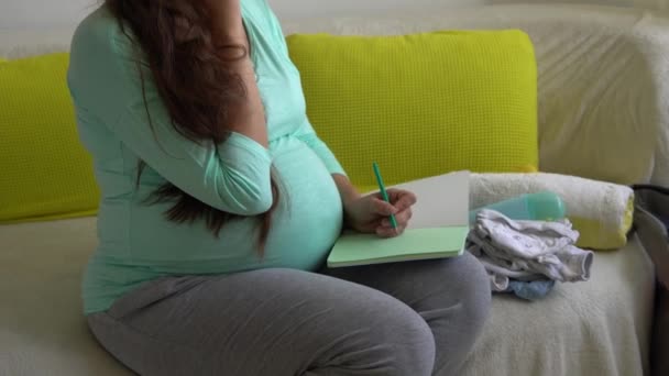 Ładna kobieta w ciąży pisze listę opakowań dla szpitala położniczego z notatnikiem Przygotowuje torby. Młody Ledy w ciąży baw się dobrze spędzając czas na kanapie w domu. Macierzyństwo, medycyna Koncepcja zdrowia i opieki — Wideo stockowe