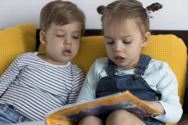 Duas crianças pré-escolares lêem um grande livro interessante de contos de fadas na cama amarela. Os irmãos pequenos leitores de gêmeos se divertem, garoto feliz em quarentena em casa. Amizade, família, conceito de educação — Fotografia de Stock