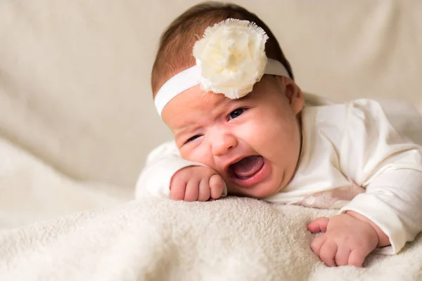 Παιδική ηλικία, μητρότητα, συναισθήματα, έννοια της μόδας - ανησυχούν θλιβερό κλάμα παχουλό μωρό με ανοιχτό το στόμα close up κοριτσάκι στο όμορφο λευκό φόρεμα floral κεφαλόδεσμο που βρίσκεται στο μαλακό ζεστό κρεβάτι στην κοιλιά στο σπίτι — Φωτογραφία Αρχείου