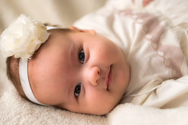 Childhood, motherhood, emotions, fashion concepts - Cute smiling happy chubby baby close up little girl in beautiful white dress flower headband on head lying on soft warm bed on her tummy at home — Stock Photo, Image