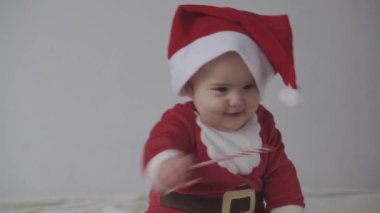 Mutlu Noeller, mutlu yıllar, bebekler, çocukluk, tatil konsepti - yakın plan gülümseyen 7 aylık Noel Baba şapkalı komik bebek, kırmızı tulum sıcak yumuşak yatağın üzerinde oturuyor tatlı Noel şekerini yalıyor