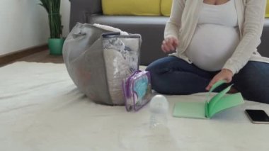 Sevimli Hamile Kadın Doğum Hastanesi için Toplanma Listesi Hazırlık Çantası Yazıyor. Hamilelikteki genç Ledy, evdeki kanepenin yanında yerde eğleniyor. Annelik, Sağlık Hizmetleri Konsepti