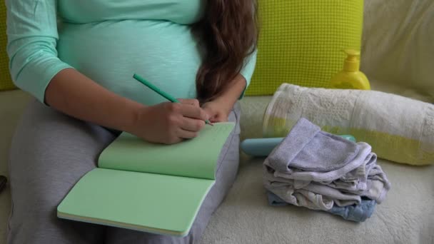 Linda mujer embarazada escribiendo lista de embalaje para el hospital de maternidad con cuaderno prepara bolsas. Joven Ledy en el embarazo divertirse pasar tiempo en el sofá en casa. Maternidad, Medicina, Salud y Atención Concepto — Vídeos de Stock