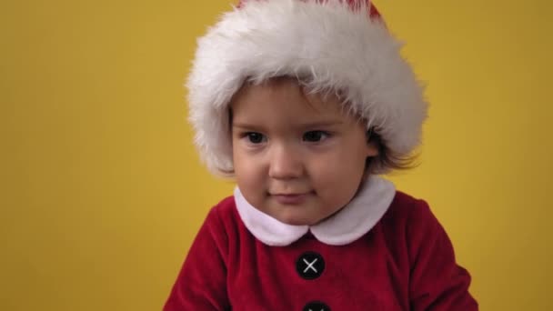 Gros plan émotion Mignon heureux joyeux joufflu tout-petit bébé fille en costume Santa regardant la caméra à fond jaune. Enfant jouant Scène de Noël Célébration d'anniversaire. Enfant amusez-vous passer du temps Nouvel An — Video