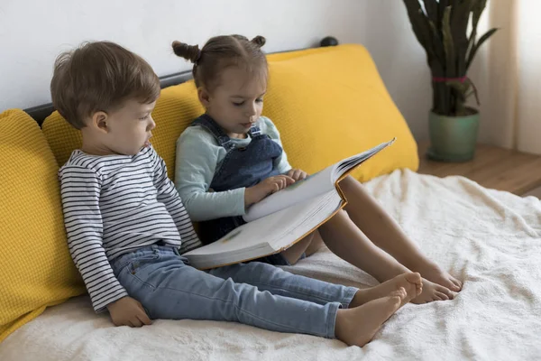 Deux enfants d'âge préscolaire, tout-petits lire grand livre intéressant de contes de fées sur lit jaune. Frères et soeurs les petits jumeaux lecteur ont plaisir, enfant heureux en quarantaine à la maison. Amitié, famille, concept d'éducation — Photo