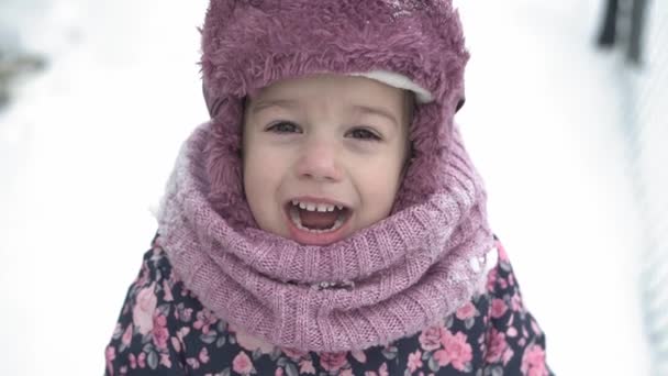 Invierno, vacaciones, juegos, conceptos familiares: primer plano slo-mo auténtica niña linda preescolar menor de 3-4 años con sombrero con orejeras mira las sonrisas de la cámara y canta en el día de invierno con clima nevado — Vídeos de Stock