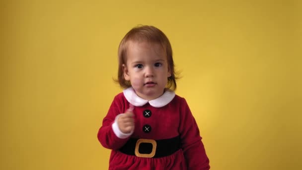 Симпатичная веселая девочка в костюме Санта-Клауса, смотрящая на камеру на желтом фоне. Детская сцена празднования дня рождения. Детский вкус есть лижет Рождество сладкие конфеты палочки Новый год — стоковое видео
