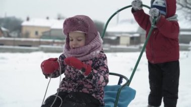 Tatil, oyun, aile kavramı - yavaş çekim otantik iki mutlu anaokulu çocuğu kardeşler şapkalar ve eldivenler takıp birbirlerini yuvarlıyorlar. Soğuk mevsimde kar yağışı, kış parkında açık havada.