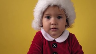 Şirin, mutlu, tombul, bebek bebek Noel Baba kostümlü, sarı arka plandaki kameraya bakıyor. Doğum gününü kutlayan Çocuk Sahne Oynuyor. Çocuk Noel oyuncağı kurabiyelerini ısırıyor Yeni yıl zamanı